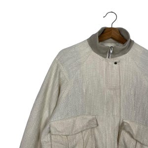May include: Off-white jacket with a textured weave, featuring a stand-up collar and two front pockets. The jacket has a zipper closure and a single button. The garment is hanging on a wooden hanger against a white background.