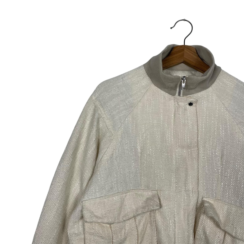 May include: Off-white jacket with a textured weave, featuring a stand-up collar and two front pockets. The jacket has a zipper closure and a single button. The garment is hanging on a wooden hanger against a white background.