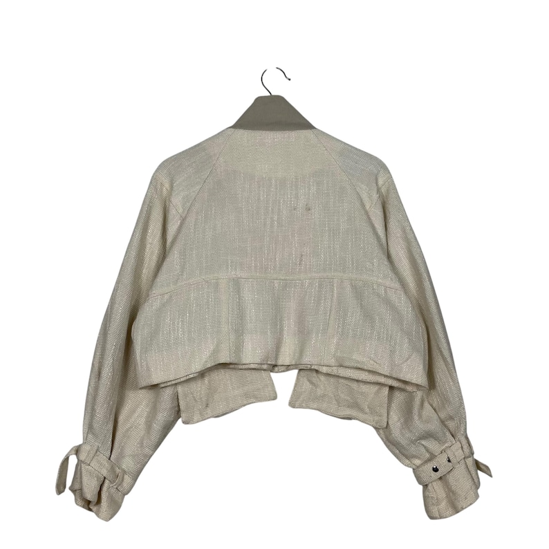 May include: A cream-colored cropped jacket with long sleeves, hanging on a hanger. The jacket features a textured fabric, a collar, and buttoned cuffs. The sleeves have tie details near the cuffs.