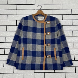 May include: A blue, grey, and white checked jacket with brown leather trim and silver clasps. The jacket is hanging on a wooden hanger. The jacket has a round collar and two pockets.