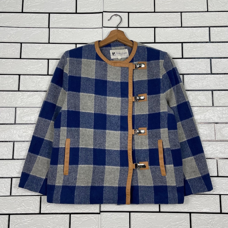 May include: A blue, grey, and white checked jacket with brown leather trim and silver clasps. The jacket is hanging on a wooden hanger. The jacket has a round collar and two pockets.