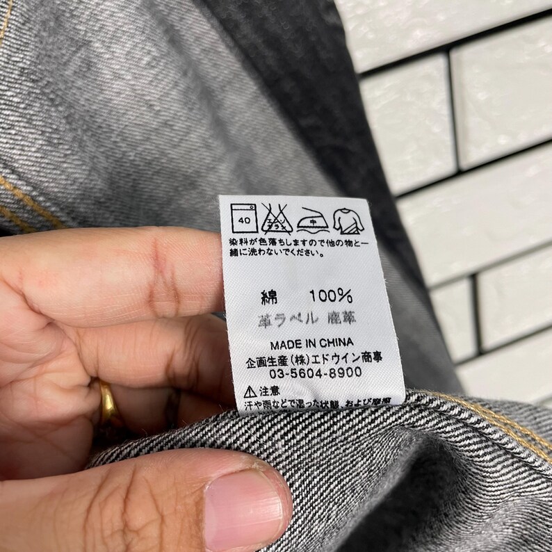 May include: Close-up of a denim garment with a white tag. The tag displays washing instructions, fabric composition (100% cotton), and the text "MADE IN CHINA". The garment is grey with yellow stitching.