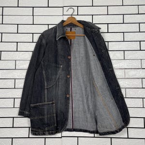 May include: A dark denim jacket with a classic collar and button-up front. The jacket has a single chest pocket and a light grey interior lining. The jacket is hanging on a wooden hanger against a white brick wall.