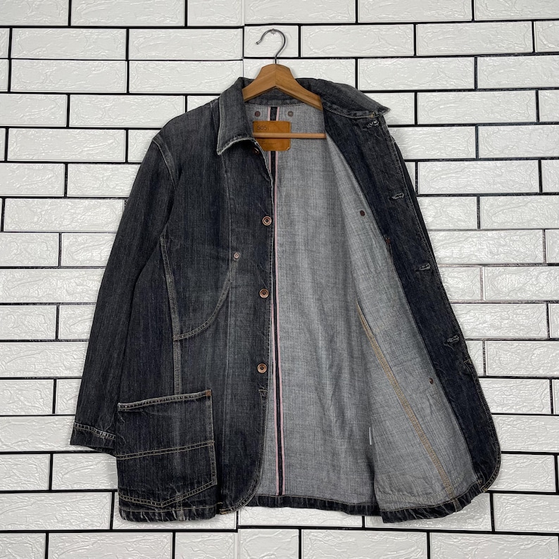 May include: A dark denim jacket with a classic collar and button-up front. The jacket has a single chest pocket and a light grey interior lining. The jacket is hanging on a wooden hanger against a white brick wall.