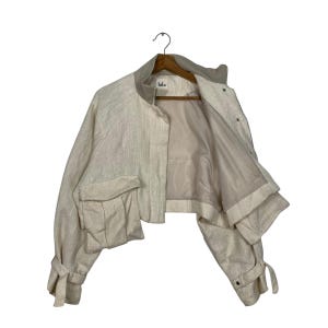 May include: A cream-colored cropped jacket with a textured weave. The jacket features a round collar, two front pockets, and tie details on the sleeves. It is hanging on a wooden hanger against a white background.