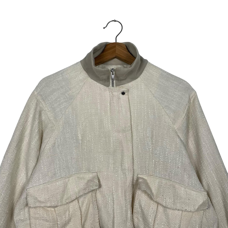 May include: Cream-colored jacket with a textured weave, featuring a high collar, a front zipper, and two front pockets. The jacket is hanging on a wooden hanger.