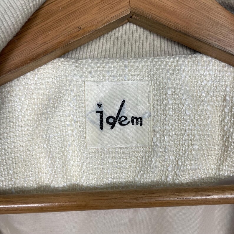May include: Close-up of a cream-colored jacket label with the brand name "idem" in black lettering. The label is rectangular and sewn onto the textured fabric of the jacket. The jacket is hanging on a wooden hanger.