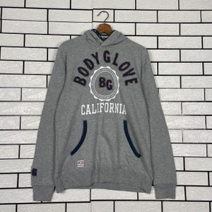 K&ouml;nnte beinhalten: Ein grauer Kapuzenpullover mit dem Text "BODY GLOVE CALIFORNIA" in Marineblau und Wei&szlig;. Der Hoodie hat eine K&auml;ngurutasche mit marineblauen Zierleisten und einen kleinen Aufn&auml;her am &Auml;rmel.