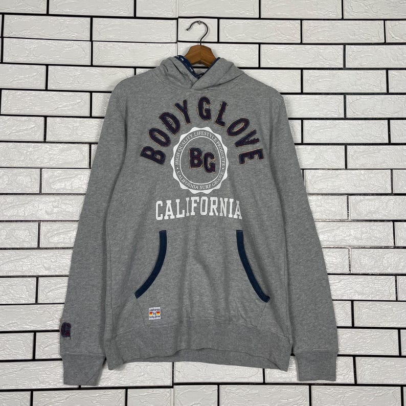K&ouml;nnte beinhalten: Ein grauer Kapuzenpullover mit dem Text "BODY GLOVE CALIFORNIA" in Marineblau und Wei&szlig;. Der Hoodie hat eine K&auml;ngurutasche mit marineblauen Zierleisten und einen kleinen Aufn&auml;her am &Auml;rmel.