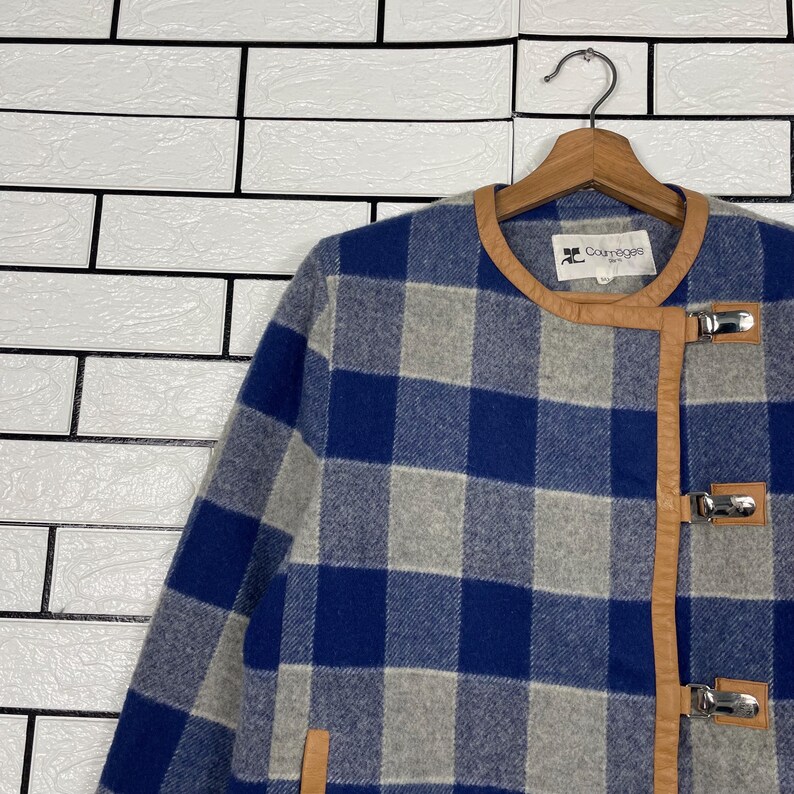 May include: A blue and grey checked jacket with brown leather trim and silver clasps. The jacket is hanging on a wooden hanger. The brand Courr&egrave;ges is visible on the collar.