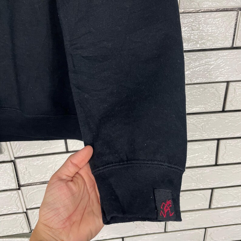 May include: Black sweatshirt with a ribbed cuff. A small, rectangular tag with a red graphic is sewn onto the cuff. The sweatshirt is made of a soft, dark fabric.