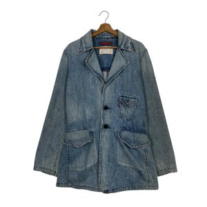 Puede incluir: Blazer de mezclilla azul claro con cuello clásico, tres botones negros y dos bolsillos delanteros. La chaqueta tiene un pequeño bolsillo en el pecho y está colgada de una percha de madera.
