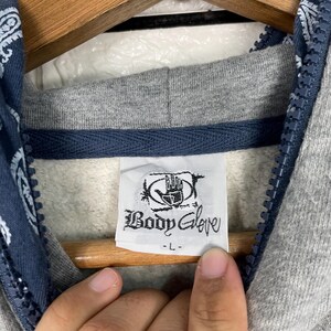 K&ouml;nnte beinhalten: Nahaufnahme eines grauen Hoodies mit blauem Paisley-Muster auf dem Kapuzenfutter. Das Etikett lautet "Body Glove". Der Hoodie hat eine graue Kapuze und einen blauen Rei&szlig;verschluss.