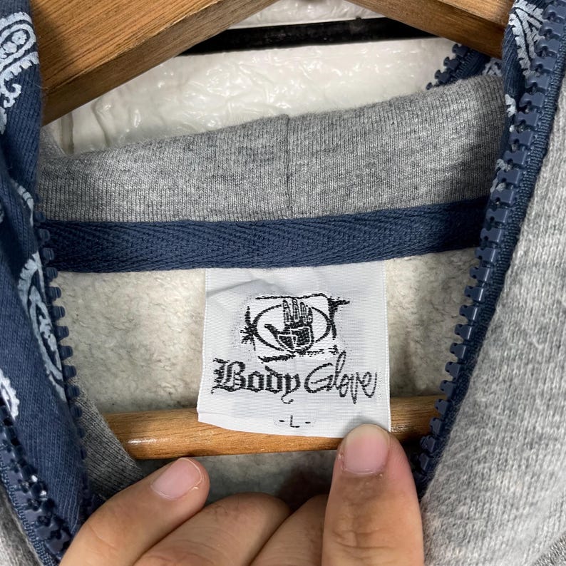 K&ouml;nnte beinhalten: Nahaufnahme eines grauen Hoodies mit blauem Paisley-Muster auf dem Kapuzenfutter. Das Etikett lautet "Body Glove". Der Hoodie hat eine graue Kapuze und einen blauen Rei&szlig;verschluss.