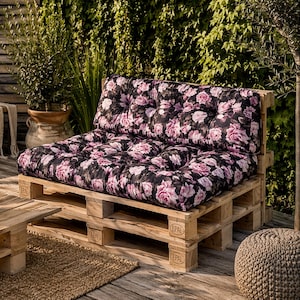 Könnte beinhalten: Ein Palettensofa mit Kissen mit rosa und lila Blumenmustern. Das Sofa besteht aus hellem Holz und befindet sich im Freien. Die Kissen haben ein getuftetes Design. Das Sofa ist Teil eines Sitzbereichs im Freien.