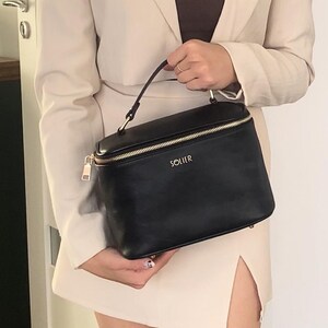 May include: A black rectangular cosmetic bag with a gold zip and handle. The bag has the word "SOLIER" in gold lettering on the front. The bag is held by a person wearing a cream-coloured blazer and skirt.