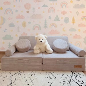 Könnte beinhalten: Ein hellgraues, modulares Spielsofa mit zwei runden Kissen und zwei Nackenrollen. Ein weißer Teddybär sitzt auf dem Sofa. Das Sofa ist aus Cordstoff gefertigt. Der Hintergrund zeigt eine Tapete mit Regenbogen, Wolken und Bäumen.
