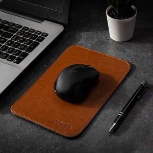 May include: A brown leather mouse pad with a black wireless mouse and a black pen. A silver laptop is in the background, and a small potted plant sits to the right. The mouse pad has the brand name "SOLIR" embossed on it.