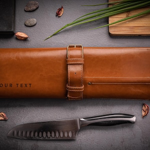 以下が含まれることがあります： 「YOUR TEXT」の文字が入った、バックル留めの茶色の革製ナイフロール。ステンレス製のシェフナイフが下に配置されています。小さな石や乾燥した葉など、装飾的な要素も含まれています。
