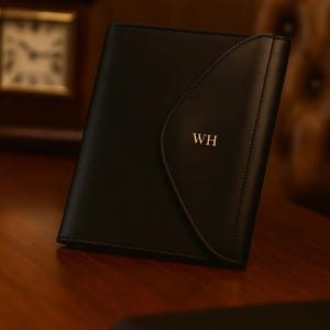 Op de afbeelding: Zwart leren folio met een gebogen flap en gestikte randen. De initialen "WH" zijn in goud op de voorkant gestempeld. De folio ligt op een houten oppervlak, met een klok en een leren stoel op de achtergrond.