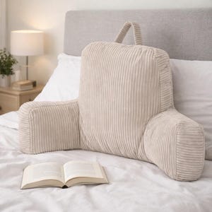 May include: A beige corduroy reading pillow with armrests and a handle. The pillow is on a bed with a book in front of it. The pillow is designed to provide back support while reading or relaxing in bed.