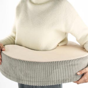 以下が含まれることがあります： ベージュとグレーのドーナツ型クッション。外側はコーデュロイ素材。クッションは曲線的なデザインで、柔らかくふわふわした表面です。人がクッションを持っています。