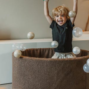 May include: A child in a black t-shirt and grey shorts is inside a brown ball pit, with clear and pearl-coloured balls in the air. The child is smiling with arms raised. The ball pit has a soft, textured exterior.