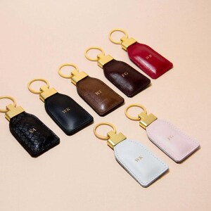 Può includere: Una collezione di portachiavi personalizzati in pelle in vari colori, tra cui nero, marrone e rosa. Ogni portachiavi presenta un anello dorato e un dettaglio metallico dorato. I portachiavi sono impressi con le iniziali.