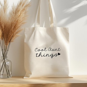 Op de afbeelding: Een crèmekleurige draagtas met de tekst "Cool Aunt things" in zwart schrift. De tas hangt aan een houten peg op een witte muur. Een heldere glazen vaas met gedroogd pampasgras staat op een houten oppervlak.