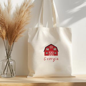 May include: Cream-colored tote bag with a red barn graphic and the word "Georgia" in red script. The bag is hanging on a wooden peg. A clear glass vase with dried pampas grass is on a wooden surface.