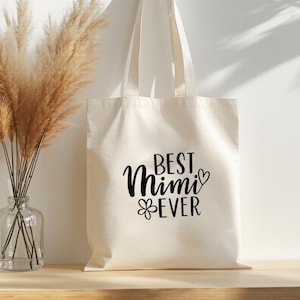 May include: Cream-colored tote bag with the words "BEST Mimi EVER" in black script. The bag is hanging on a wooden peg. A vase of dried pampas grass sits on a wooden surface to the left.