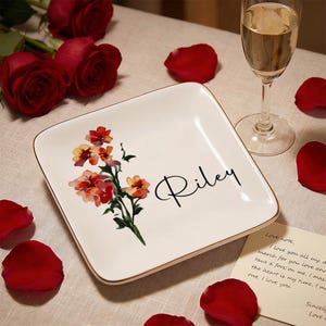 May include: A square, white ceramic plate with a gold rim, featuring a floral design in red and orange hues. The name "Riley" is written in black script. Red rose petals and a glass of champagne are in the background.