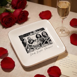 Może przedstawiać: Biały kwadratowy ceramiczny talerz ze złotą obwódką, z czarno-białym zdjęciem czterech kobiet. Czerwone płatki róż i róże otaczają talerz. Obecny jest również kieliszek szampana i odręczna notatka.