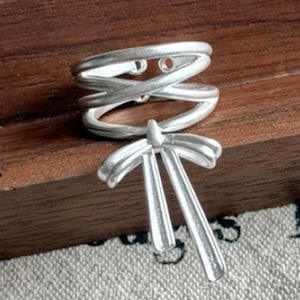 Può includere: Un anello color argento con un design intricato. L'anello presenta una sezione superiore a doppio incrocio e un elemento inferiore stilizzato a forma di ventaglio. L'anello è esposto su una superficie di legno.