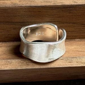Pode incluir: Um anel de prata com acabamento escovado. O anel tem uma banda larga com um design curvo e aberto. O interior do anel é estampado com o número "925". O anel é exibido em uma superfície de madeira.