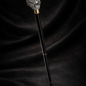 May include: A black walking cane featuring a silver skull-shaped handle. The skull has detailed facial features and a metallic finish. The cane has gold accents and a sleek, polished appearance.