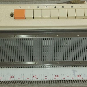 Puede incluir: Máquina de tejer Brother KH 710, de color crema, con botones blancos y naranjas. Cuenta con un indicador con números y un contador de filas. El texto "Brother KH 710" es visible.