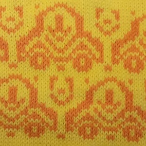 May include: Close-up of a knitted fabric with a repeating pattern of orange cars on a yellow background. The cars have a simple, stylized design. The fabric appears to be soft and textured, with visible knit stitches.