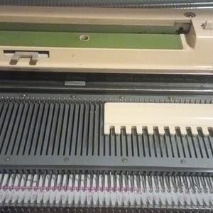 Puede incluir: Primer plano de una máquina de tejer vintage. La máquina tiene un cuerpo color crema con un acento verde. Las agujas están dispuestas en filas y una pieza blanca en forma de peine está en su lugar. La máquina está diseñada para crear tejido de punto.