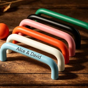 May include: A collection of colorful cabinet pulls in various shades, including green, black, pink, orange, white, and light blue. The light blue pull has the text "Alice & David". Also included are two orange knobs. The hardware is arranged on a wooden surface.