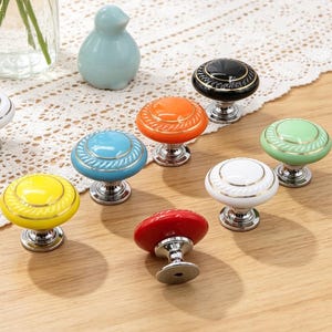 May include: A collection of colorful ceramic cabinet knobs with silver bases. The knobs are in various colors, including white, yellow, blue, orange, green, red, and black. Each knob has a decorative design and is arranged on a wooden surface.