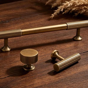 May include: A set of antique brass cabinet hardware, including a long bar pull, a round knob, and a smaller pull. Each piece features a ribbed design and a warm, golden finish. The hardware is displayed on a dark wood surface.