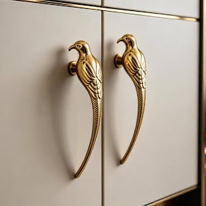 May include: Two gold-colored bird-shaped cabinet handles. The handles are mounted on a light beige cabinet door. The bird handles have detailed feather and beak designs. The cabinet has a gold trim.