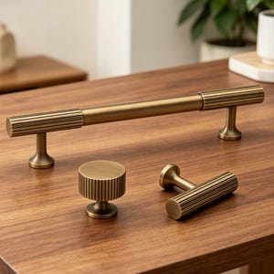 May include: A collection of brass cabinet hardware, including a long bar pull, a round knob, and a smaller pull. The hardware features a ribbed design and is displayed on a wooden surface.