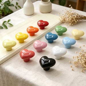 May include: A collection of heart-shaped cabinet knobs in various colors, including red, orange, yellow, green, blue, pink, white, and black. The knobs are arranged on a light-colored wooden board, with a neutral background.
