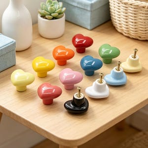 May include: A collection of heart-shaped cabinet knobs in various colors, including red, orange, yellow, pink, blue, green, white, and black. Each knob has a gold-colored screw for installation. The knobs are arranged on a light-colored wooden surface.