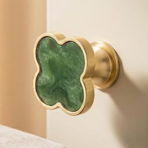 May include: A decorative cabinet knob with a four-leaf clover design. The knob features a gold-colored metal base and a green, marbled-effect center. The knob is mounted on a cream-colored cabinet door.