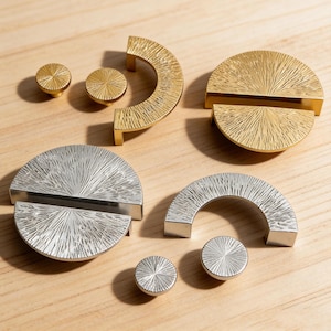 Könnte beinhalten: Eine Sammlung von gold- und silberfarbenen Schrankbeschlägen, darunter Knöpfe und Griffe. Die Beschläge weisen ein strukturiertes, radiales Design auf. Die Teile sind auf einer hellen Holzoberfläche angeordnet und zeigen ihre unterschiedlichen Formen und Oberflächen.
