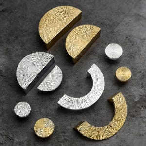 May include: A selection of gold and silver cabinet hardware, including half-circle pulls, round knobs, and curved handles. The hardware has a textured, sunburst design. The pieces are metal and are arranged on a dark surface.