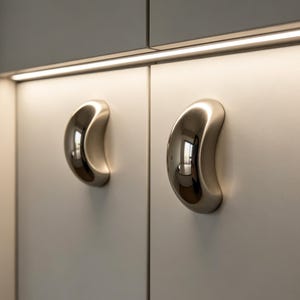 May include: Close-up of two cabinet doors with brushed nickel crescent-shaped handles. The handles are mounted on a light gray cabinet door. The cabinet doors are positioned side by side, with a warm light strip above.